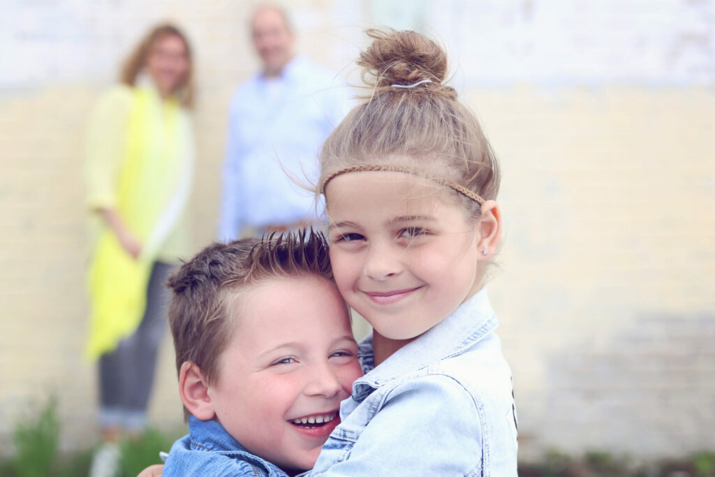 Broer en zus knuffelen lachend met hun ouders op de achtergrond