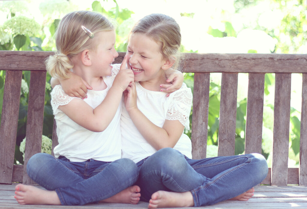 Twee zusjes zitten knus tegen elkaar op een houten vlonder, kinderfoto buiten