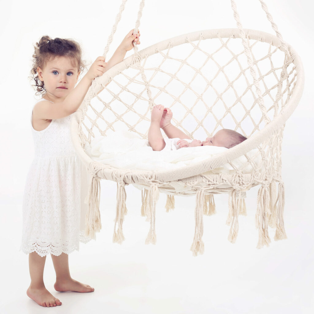 Peuter in wit jurkje bij een macramé hangstoel met newborn