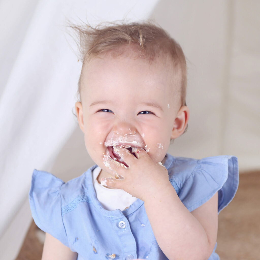 Lachend babymeisje in lichtblauw jurkje eet taart, cakesmash
