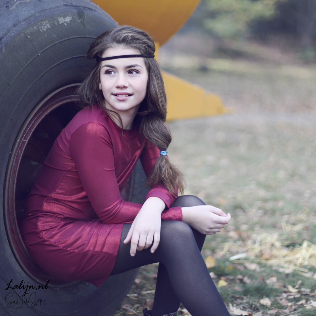 Meisje in bordeauxrode jurk poseert bij een betonnen buis, kinderfotografie