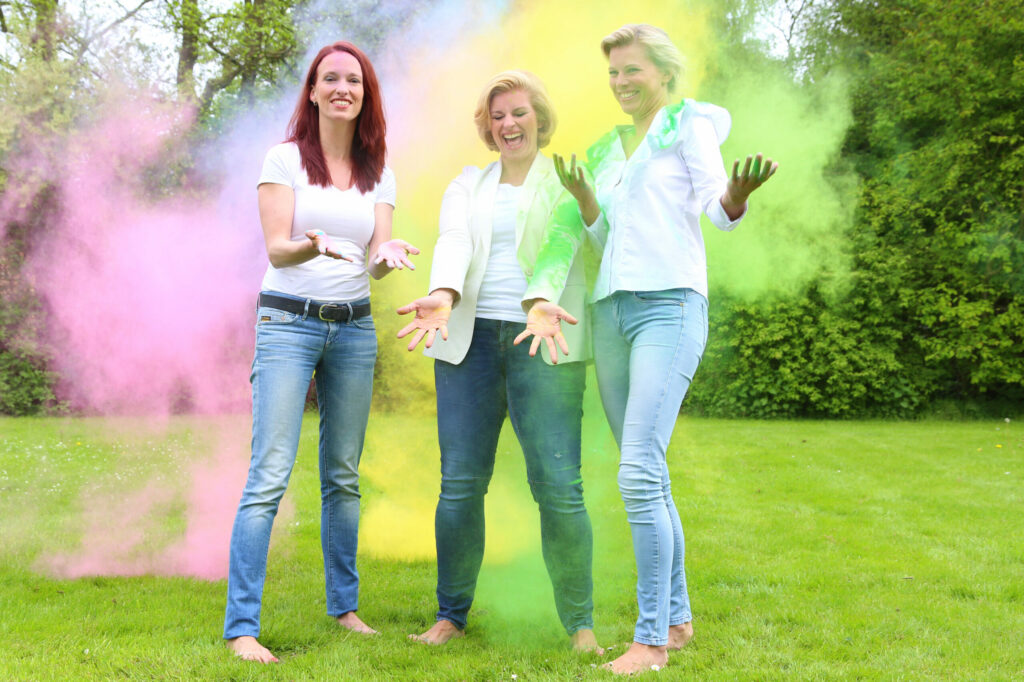 Drie vrouwen staan lachend in roze en gele poederwolken op het gras