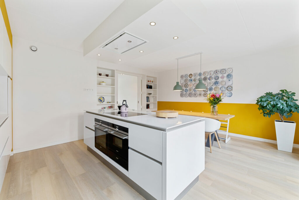 Witte keuken met kookeiland, eettafel en gele accentwand