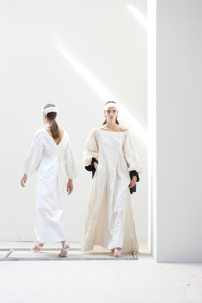 Twee modellen in witte outfits lopen door een lichte hal, modeshow