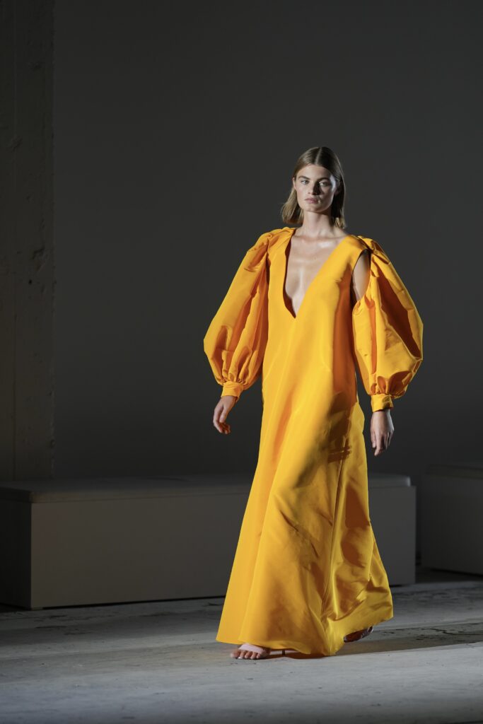 Model in okergele jurk met pofmouwen op de catwalk