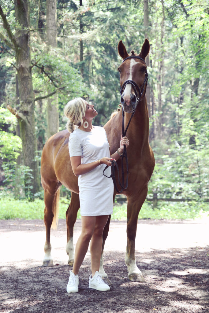 Vrouw in witte jurk naast haar bruine paard op een bospad, paardenshoot