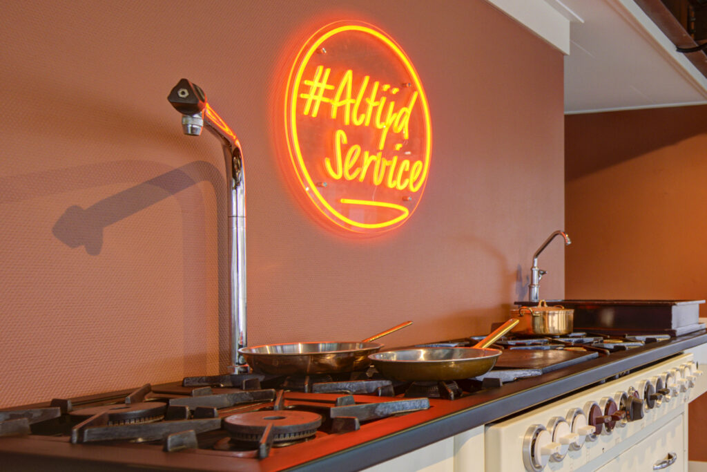 Bedrijfskeuken met neonbord '#Altijd service' aan een oranje wand